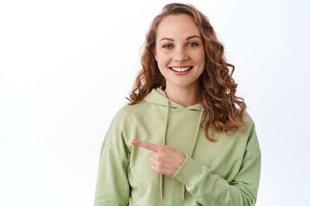 Smiling pretty girl with curly fair hair, pointing finger aside, showing copyspace promotional text, standing in green hoodie against white wall