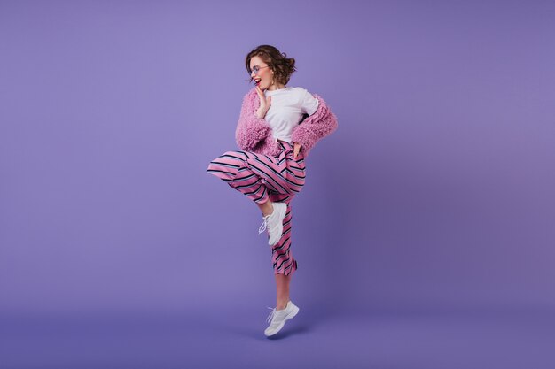 Smiling pretty girl with wavy hairstyle standing on one leg on purple wall.  cheerful brunette female model dancing in white sneakers.