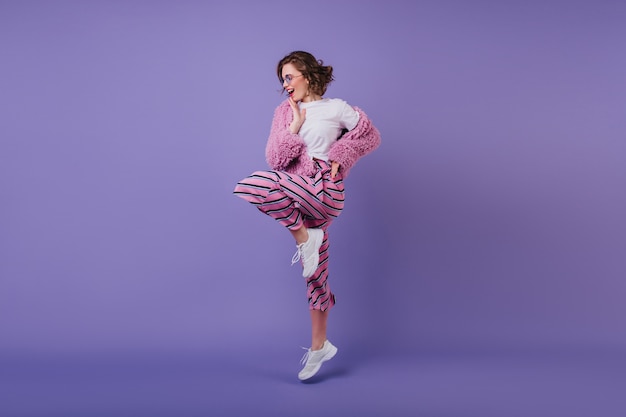 紫色の壁に片足で立っている波状の髪型のかわいい女の子の笑顔。白いスニーカーで踊る陽気なブルネットの女性モデル。
