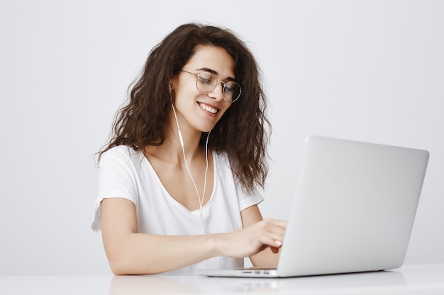 Улыбающаяся красивая девушка работает с ноутбуком и слушает музыку в наушниках