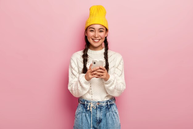 笑顔のかわいいミレニアル世代の女の子は、ワイヤレスインターネットに接続された最新の携帯電話を使用し、画像をダウンロードし、メールボックスをチェックし、黄色い帽子をかぶっています