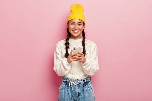 Smiling pretty millennial girl uses modern mobile phone, connected to wireless internet, downloads images, checks email box, wears yellow hat
