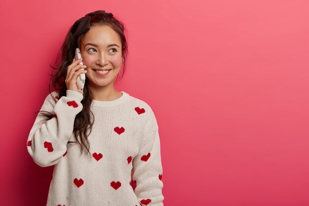 Smiling pretty mixed race female talks on cellphone, discusses what happened during day with mother, has happy look, wears white jumper