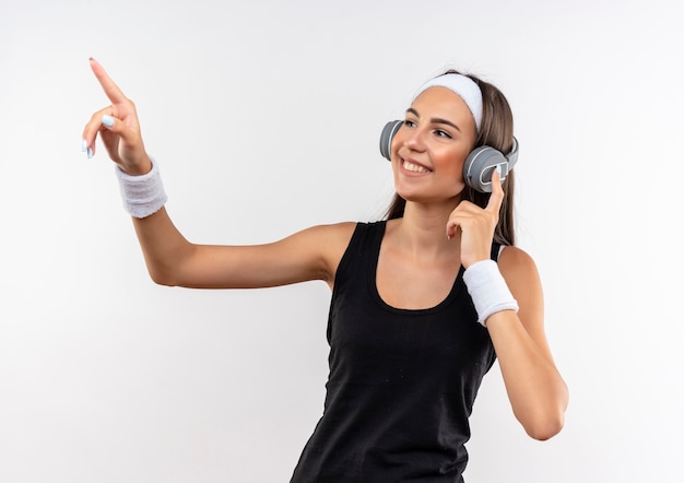 Smiling pretty sporty girl wearing headband and wristband and headphones pointing at side isolated on white space