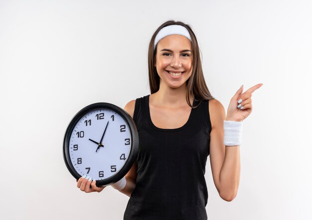 Smiling pretty sporty girl wearing headband and wristband holding clock and pointing at side isolated on white space