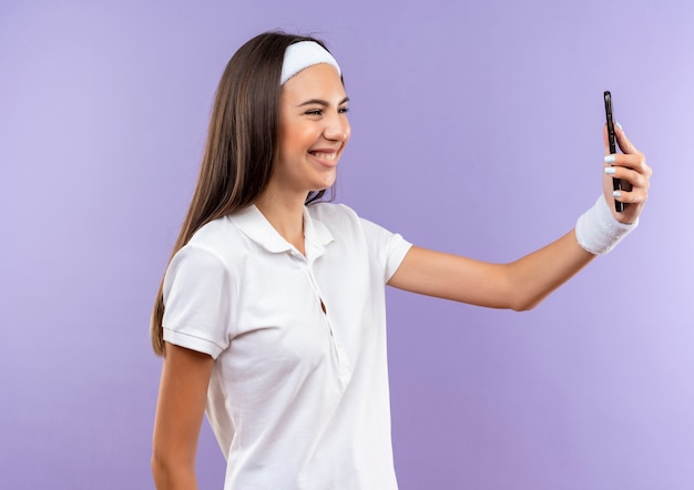 Smiling pretty sporty girl wearing headband and wristband holding and looking at mobile phone isolated on purple space