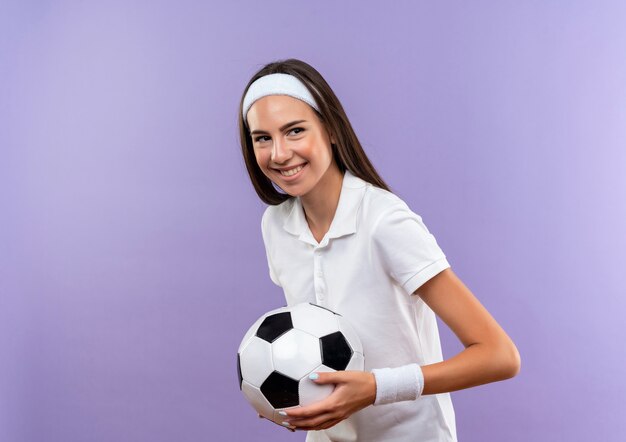 Улыбающаяся симпатичная спортивная девушка с головной повязкой и браслетом держит футбольный мяч на фиолетовом пространстве