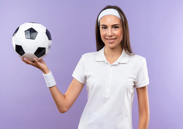 Smiling pretty sporty girl wearing headband and wristband holding soccer ball isolated on purple space