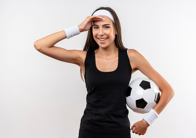 Smiling pretty sporty girl wearing headband and wristband holding soccer ball and putting hand on forehead isolated on white space