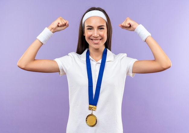 Улыбающаяся симпатичная спортивная девушка с обручем и браслетом и медалью жестко жестикулирует на фиолетовом пространстве