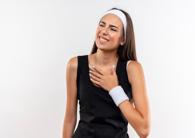 Smiling pretty sporty girl wearing headband and wristband putting hand on chest looking at side isolated on white space 