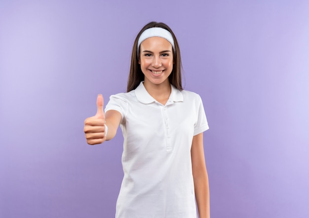 Smiling pretty sporty girl wearing headband and wristband showing thumb up isolated on purple space