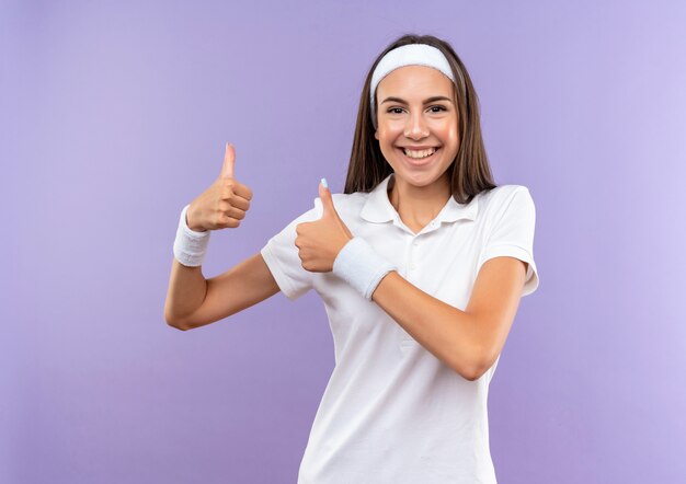 Smiling pretty sporty girl wearing headband and wristband showing thumbs up isolated on purple space