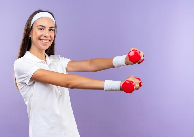 Smiling pretty sporty girl wearing headband and wristband stretching out hands isolated on purple space