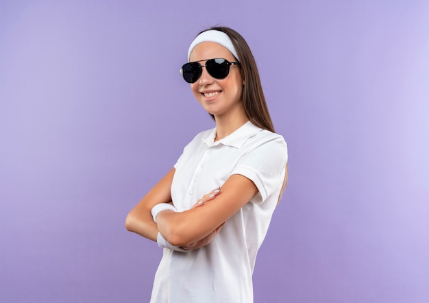 Smiling pretty sporty girl wearing headband and wristband and sunglasses standing with closed posture isolated on purple space