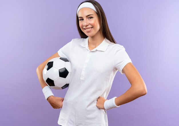 Smiling pretty sporty girl wearing headband and wristband with soccer ball putting hands on waist isolated on purple space