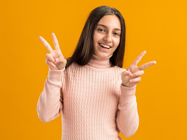 Улыбающаяся симпатичная девочка-подросток смотрит вперед, делая знак мира обеими руками, изолированными на оранжевой стене с копией пространства