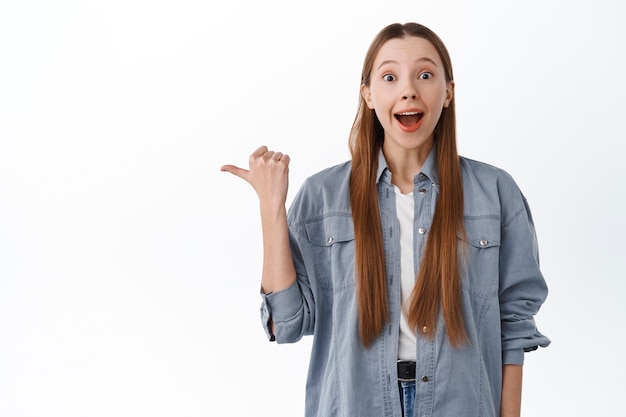 Smiling pretty teenage girl with long healthy hair smiling, pointing thumb aside at promo text, showing advertisement copy space, white wall