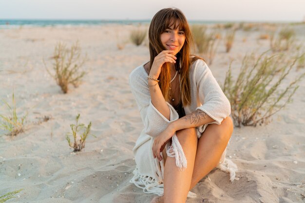 모래에 앉아 세련된 목걸이와 보헤미안 여름 복장에 웃는 예쁜 여자