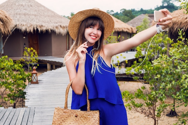 Улыбается красивая женщина, делая автопортрет по мобильному телефону на своем тропическом отдыхе в Таиланде. Летняя ярко-синяя одежда ^ соломенная модная шляпа и сумка. Красные губы. Хорошее настроение.