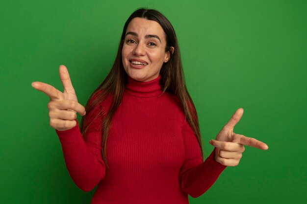 Smiling pretty woman points at front with two hands isolated on green wall