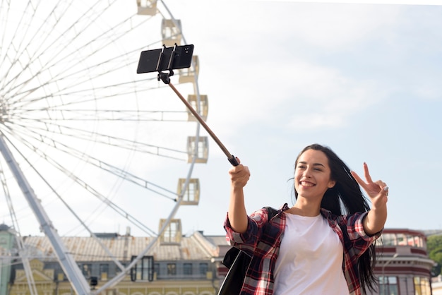 Усмехаясь милая женщина принимая selfie с показывать жест победы стоя около колеса обозрения