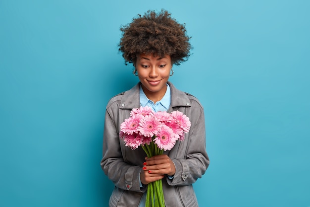 La donna graziosa sorridente con capelli ricci naturali osserva felicemente il mazzo della margherita del gerbera