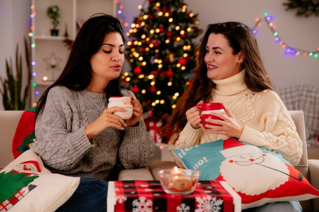 笑顔のかわいい若い女の子はカップを保持し、目を閉じて肘掛け椅子に座って、家でクリスマスの時間を楽しんでカップを保持している彼女の喜んでいる友人を見ます