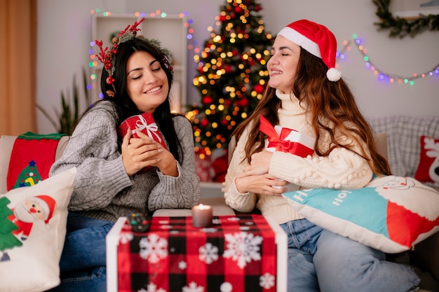 Smiling pretty young girl with santa hat holds gift box and looks at her pleased friend with holly wreath sitting on armchairs and enjoying christmas time at home