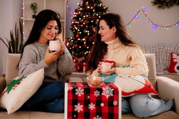 笑顔のかわいい若い女の子が肘掛け椅子に座って、家でクリスマスの時間を楽しんでカップを持って見て