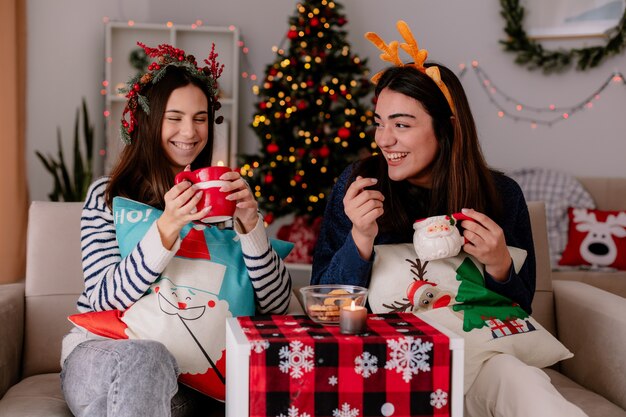 Smiling pretty young girls with holly wreath and reindeer headband hold cups sitting on armchairs and enjoying christmas time at home