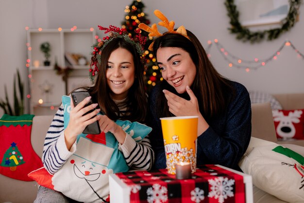 smiling pretty young girls with holly wreath and reindeer headband look at phone sitting on armchairs and enjoying christmas time at home