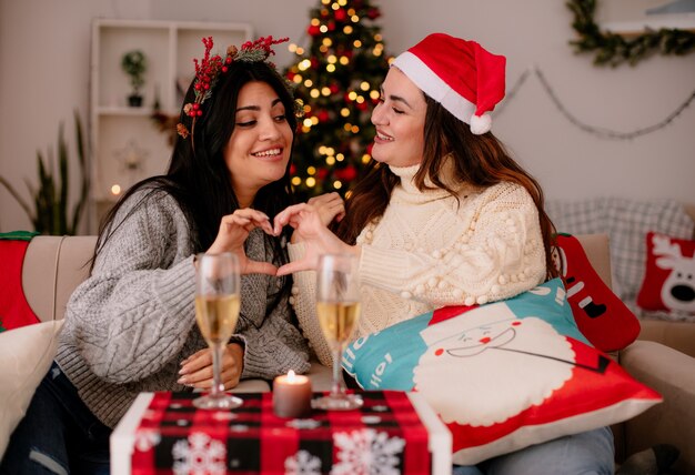 Smiling pretty young girls with santa hat gesturing heart sign together sitting on armchairs and enjoying christmas time at home