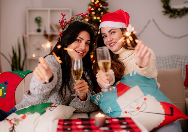 Smiling pretty young girls with santa hat hold glasses of champagne and sparklers sitting on armchairs and enjoying christmas time at home