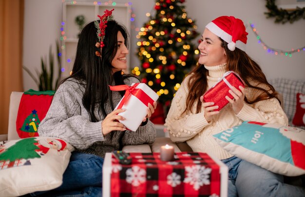 Smiling pretty young girls with santa hat and holly wreath hold gift boxes and look at each other sitting on armchairs and enjoying christmas time at home
