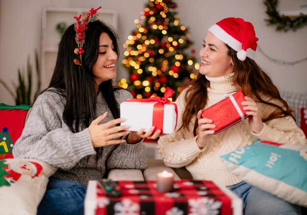 Smiling pretty young girls with santa hat and holly wreath hold gift boxes sitting on armchairs and enjoying christmas time at home