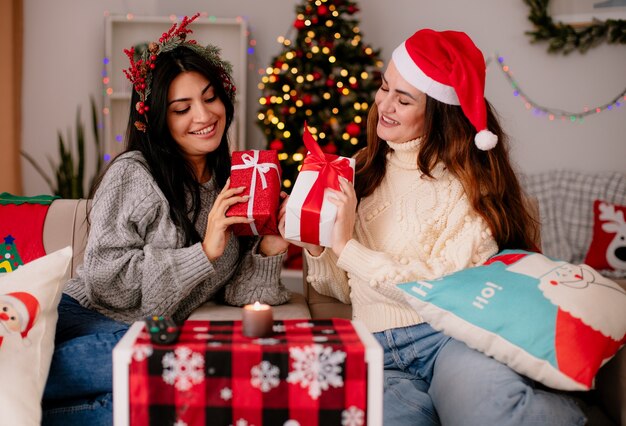 улыбающиеся симпатичные молодые девушки в шляпе санта-клауса и венок из холли держатся и смотрят на свои подарочные коробки, сидя на креслах и наслаждаясь Рождеством дома