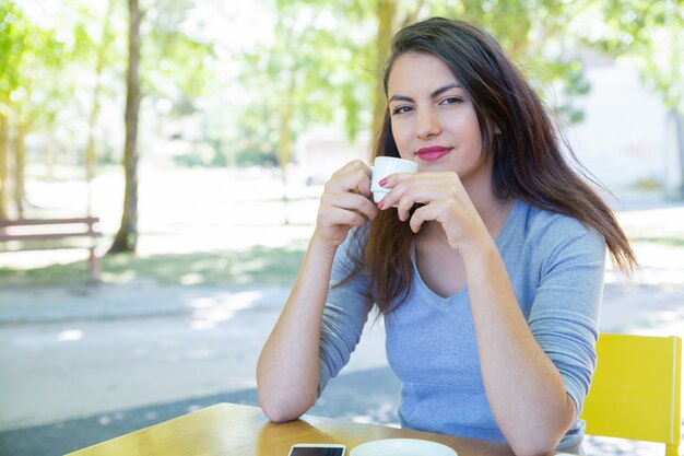 Smiling pretty young lady drinking coffee at cafe table in park