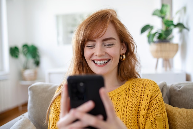 웃고 있는 예쁜 젊은 여성은 소셜 미디어와 인터넷 서핑을 위해 모바일 스마트 폰을 사용하는 동안 거실 소파에서 휴식을 취합니다.