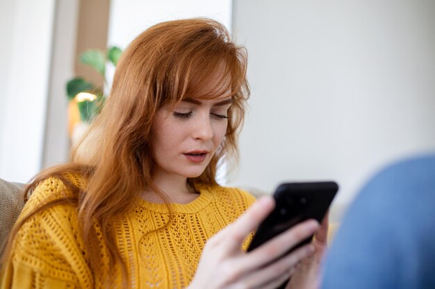 La bella giovane donna sorridente si rilassa sul divano nel suo soggiorno mentre usa il suo smartphone mobile per i social media e naviga in internet