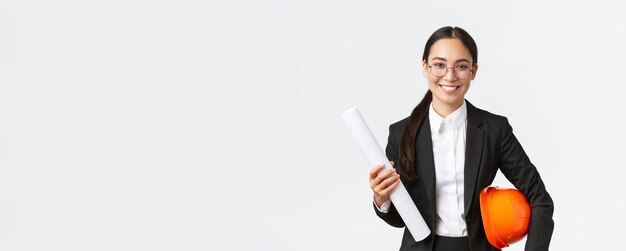 Smiling professional asian female architect in business suit and glasses carry blueprints of home design and safety helmet engineer introduce her project enter construction area white background