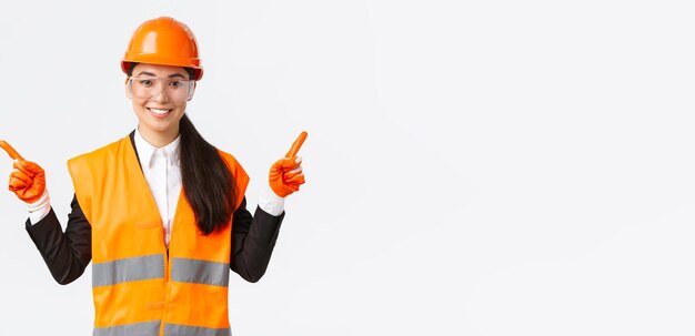 Smiling professional asian female builder engineer in safety helmet protective glasses pointing fingers sideways showing left and right project introduce construction projects white background