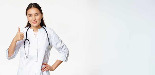 Smiling professional doctor in medical uniform shows thumbs up pleased asian female physician confir