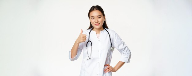 Улыбающийся профессиональный врач в медицинской форме показывает палец вверх Довольная азиатская женщина-врач подтверждает что-то, рекомендуя продукт на белом фоне