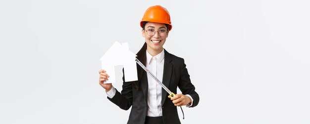 Smiling professional female asian engineer architect in helmet and business suit showing house maket and tape measure ready starting home renovation construction works white background