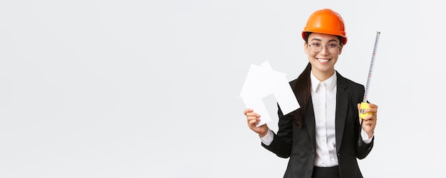 Smiling professional female asian engineer architect in helmet and business suit showing house maket and tape measure ready starting home renovation construction works white background