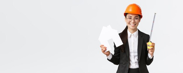 Улыбающаяся профессиональная азиатская женщина-архитектор в шлеме и деловом костюме, показывающая домашний макияж и рулетку, готовую к началу ремонта дома, строительные работы на белом фоне