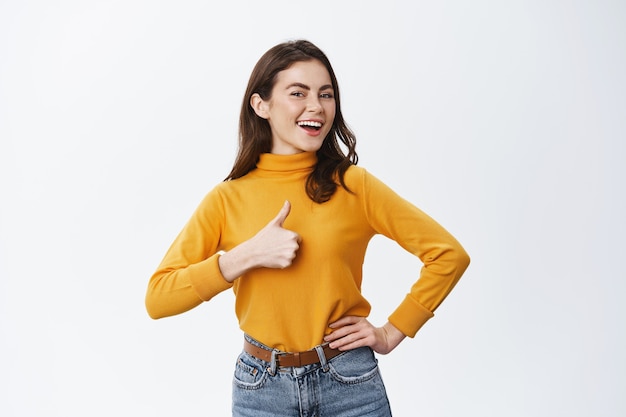 Smiling proud woman showing thumb up and say yes, nod to agree or approve, praising good work or nice choice, standing against white wall