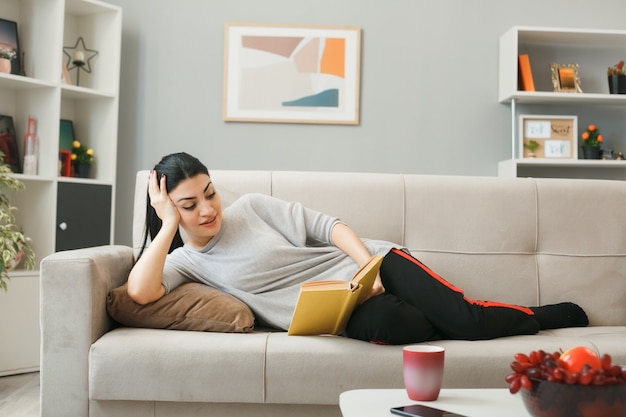 Free photo smiling putting hand on cheek young girl reading book lying on sofa behind coffee table in living room