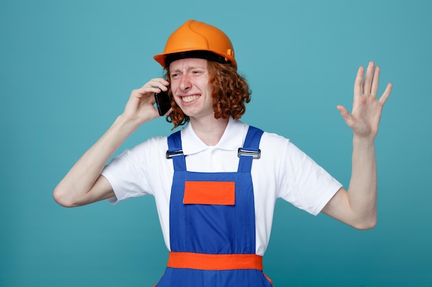 Smiling raising hand young builder man in uniform speak on the phone isolated on blue background